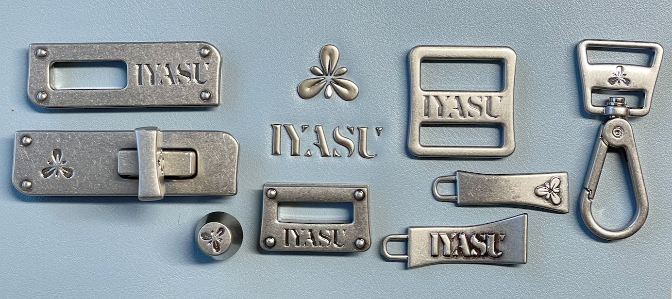 Custom alloy hardware pieces with logos and motifs designed for IYASU bags are shown alongside a black bag featuring these branded elements.